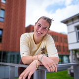 A smiling young woman is leaning on a railing with buildings in the background